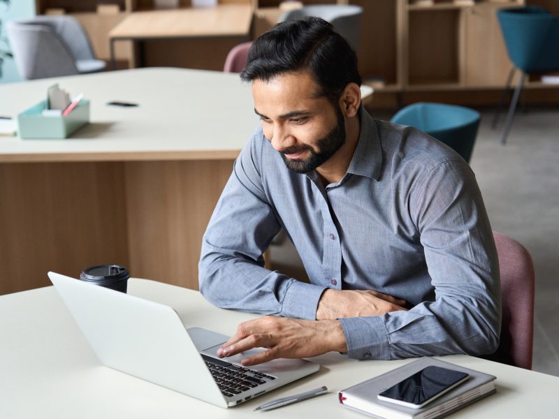 Smiling indian business man working with laptop and cellphone in coworking.